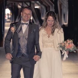 Ein Brautpaar geht auf einem rustikalen, hölzernen Brücke. Der Mann trägt einen dunklen Anzug und die Frau ein weißes Brautkleid.