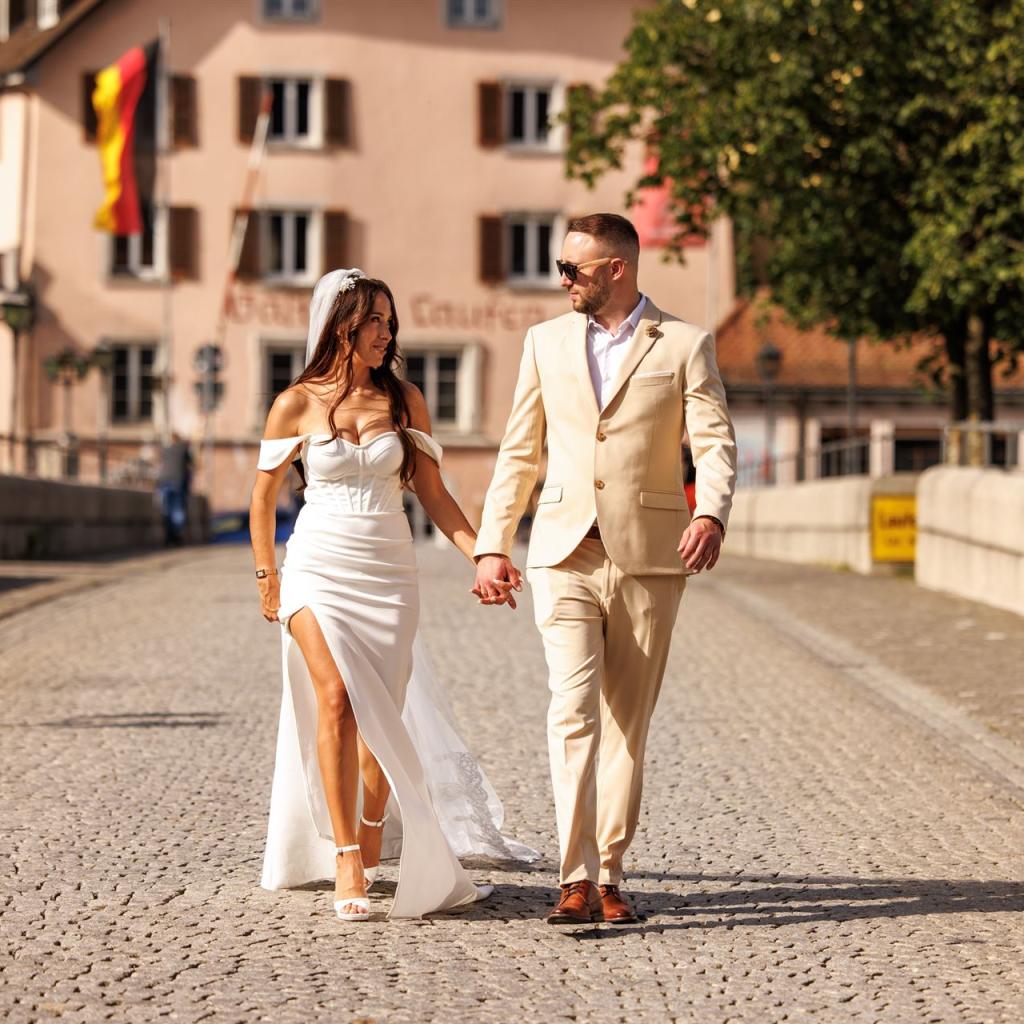 Ein junges Paar, das Hand in Hand durch eine sonnige, gepflasterte Straße in einer historischen Stadt spaziert.