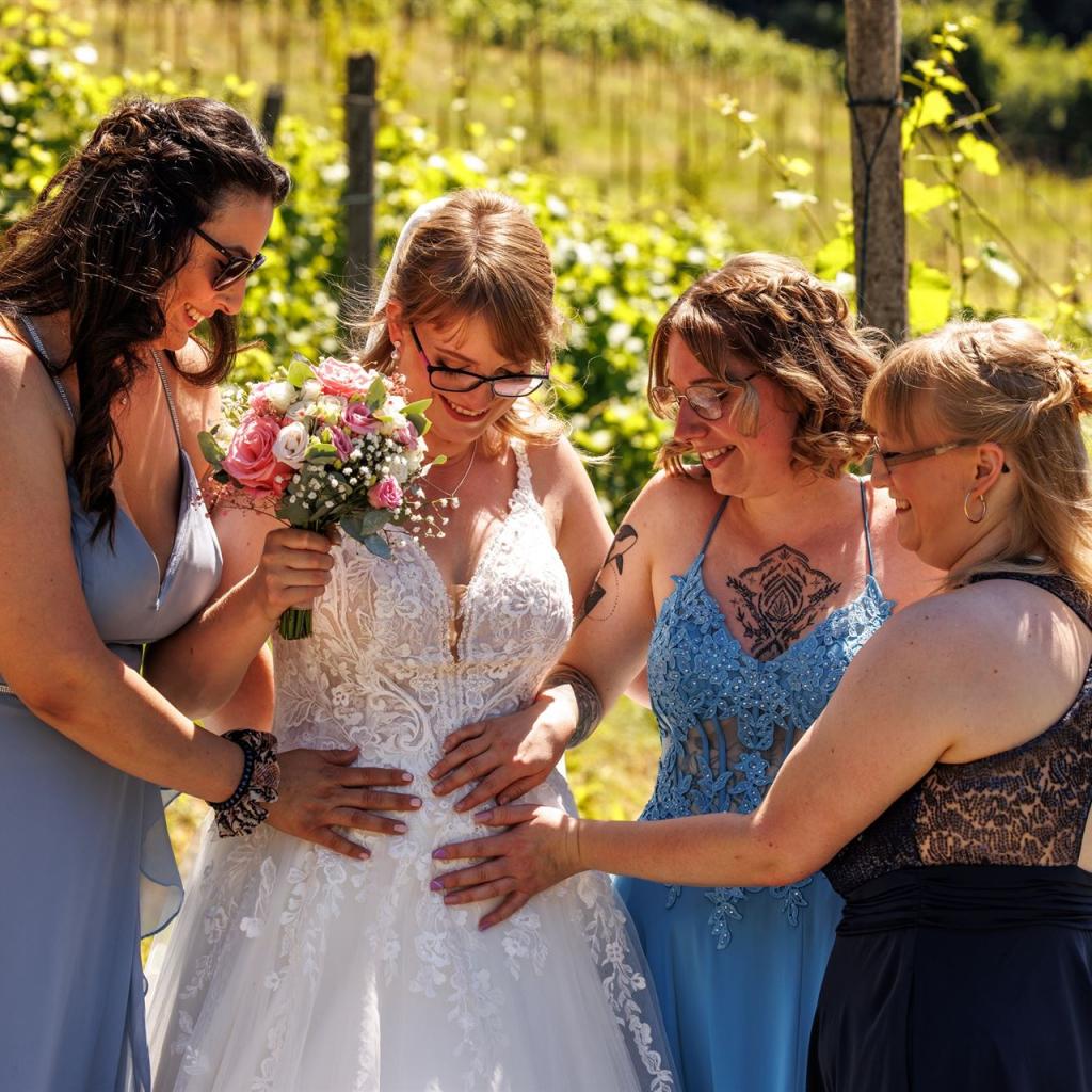 Eine Gruppe von vier Frauen in festlicher Kleidung steht lachend in einem Weinberg mit viel Grün.