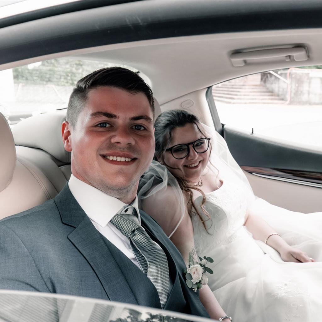 Ein frisch verheiratetes Paar sitzt glücklich in einem Auto, die Frau trägt ein Brautkleid und lächelt.